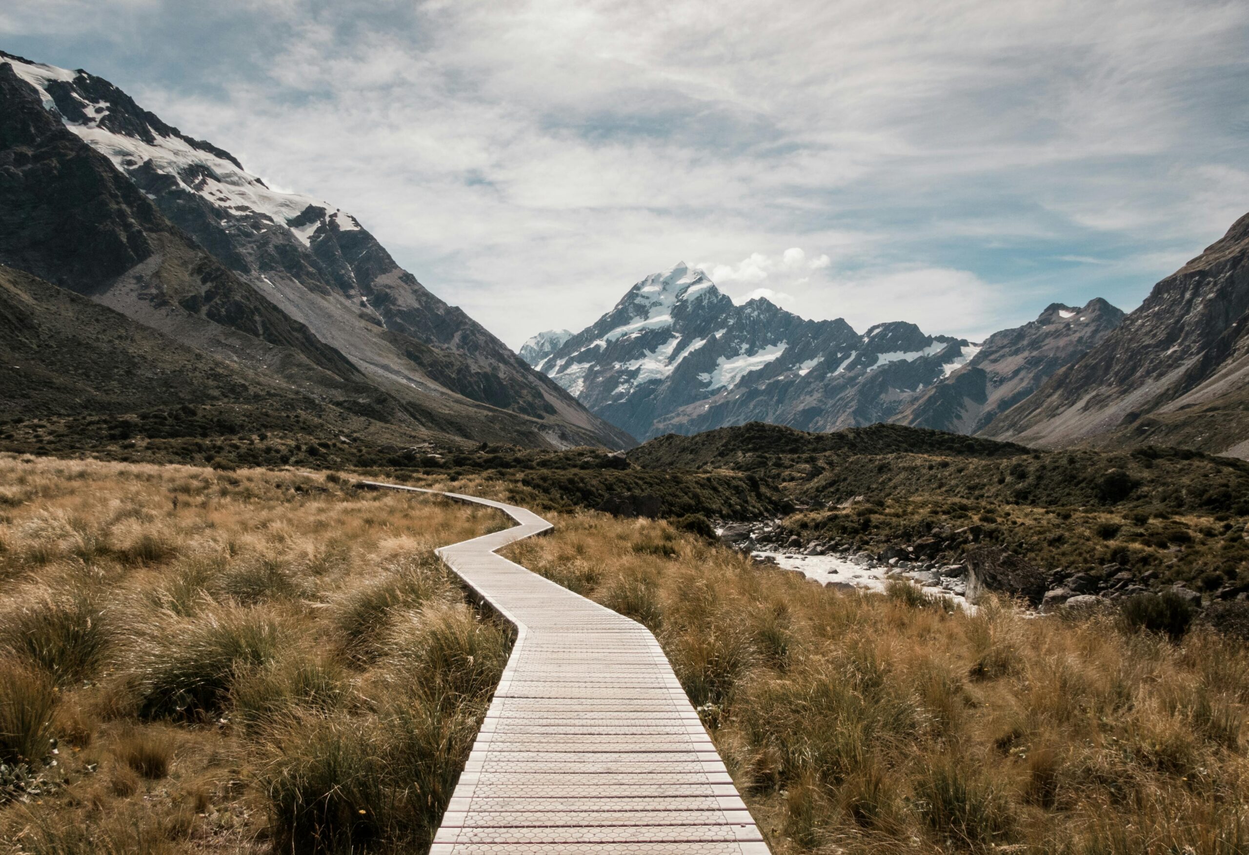 Wooden walkway leads through rugged landscape towards majestic snow-capped mountains.